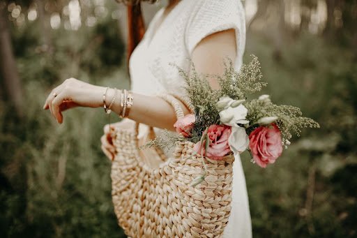 A woman wearing a white dress holds a woven bag adorned with greenery and pink flowers in an outdoor setting.