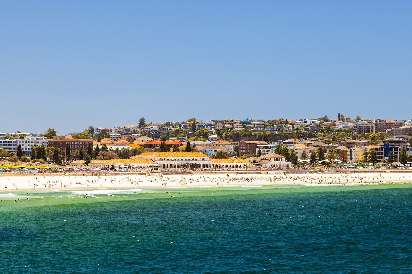  A sweeping view of Bondi Beach, with golden sand, turquoise waters and a lively beachfront lined with beach enthusiasts, caf&eacute;s, restaurants and residential buildings.
