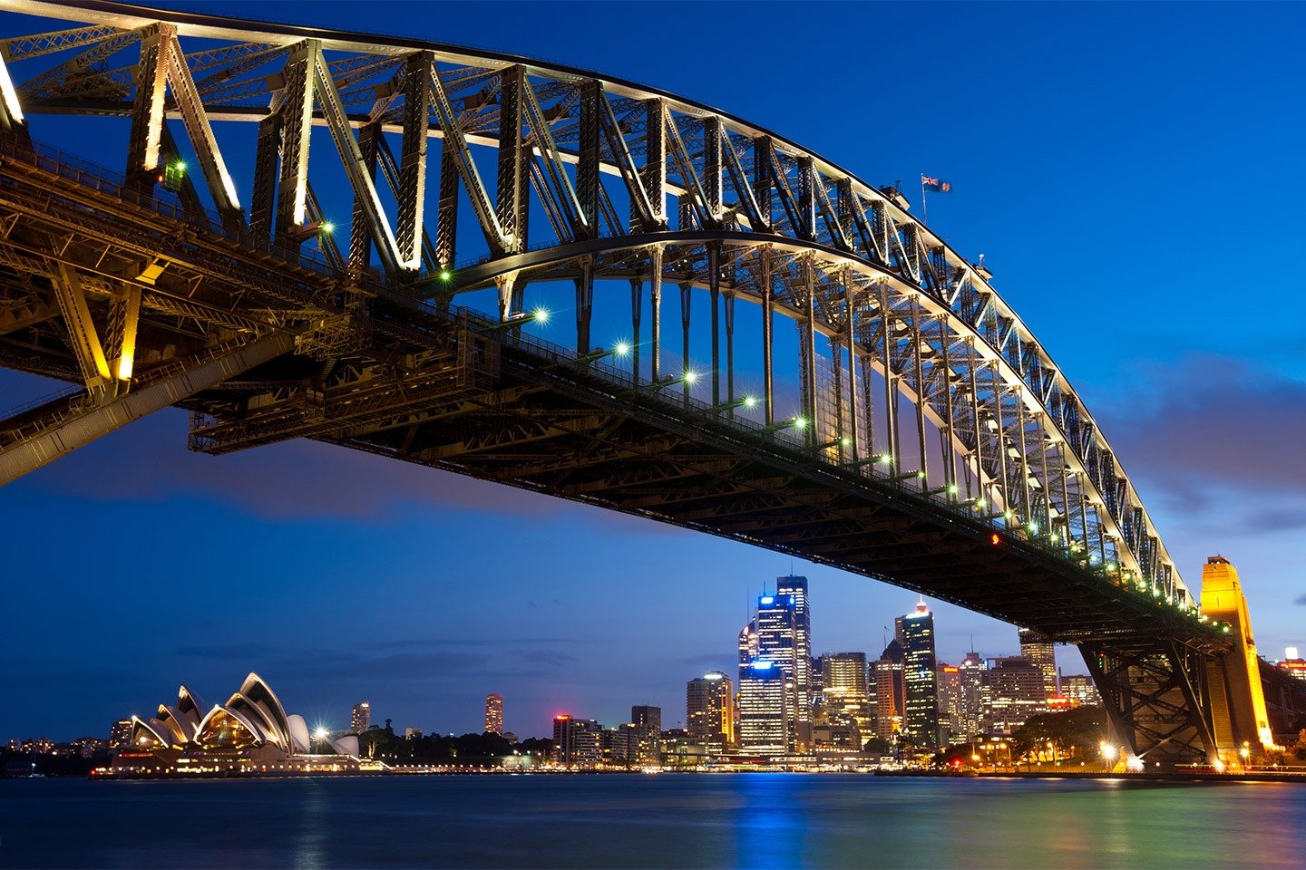 The Sydney Harbour Bridge illuminated at dusk, with the Sydney Opera House and the city's glowing skyline visible along the waterfront.