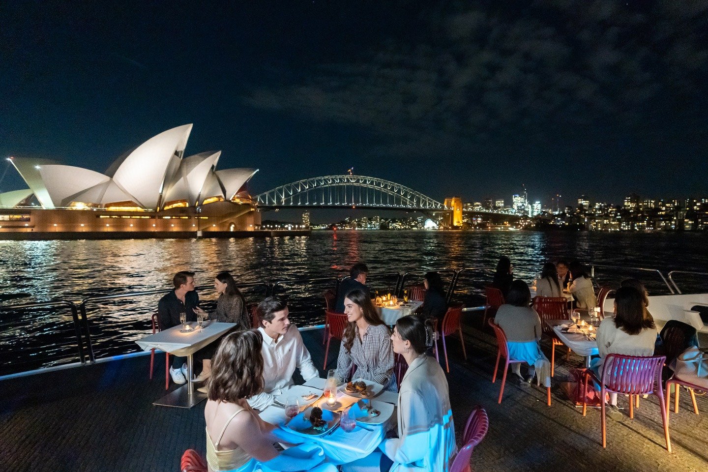 Guests dining on a Sydney Harbour dinner cruise, with the illuminated Sydney Opera House and Harbour Bridge in the background.
